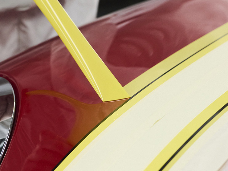 Jemand klebt einen Streifen gelbes Abdeckband auf die Oberfläche eines roten Autos, um es für Lackier- oder Polierarbeiten vorzubereiten.